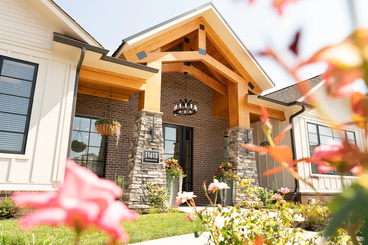 The entrance features a wooden archway illuminated by a chandelier, with a brick facade and colorful flower planters. Children play in the garden, suggesting a family-friendly and welcoming atmosphere, while adults gather under the chandelier in the evening.