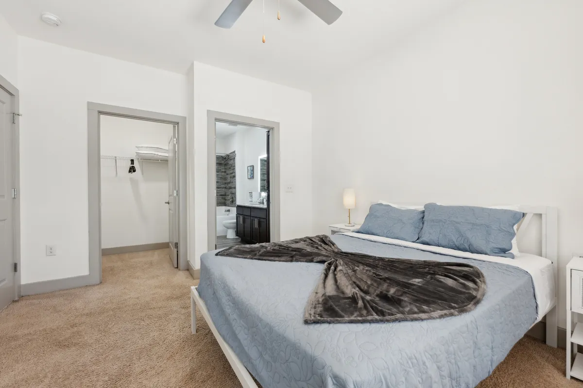 A cozy bedroom featuring blue bedding, a bedside lamp, an open closet, and a visible entrance to the bathroom. The room appears welcoming and organized, with soft lighting enhancing its comfort.