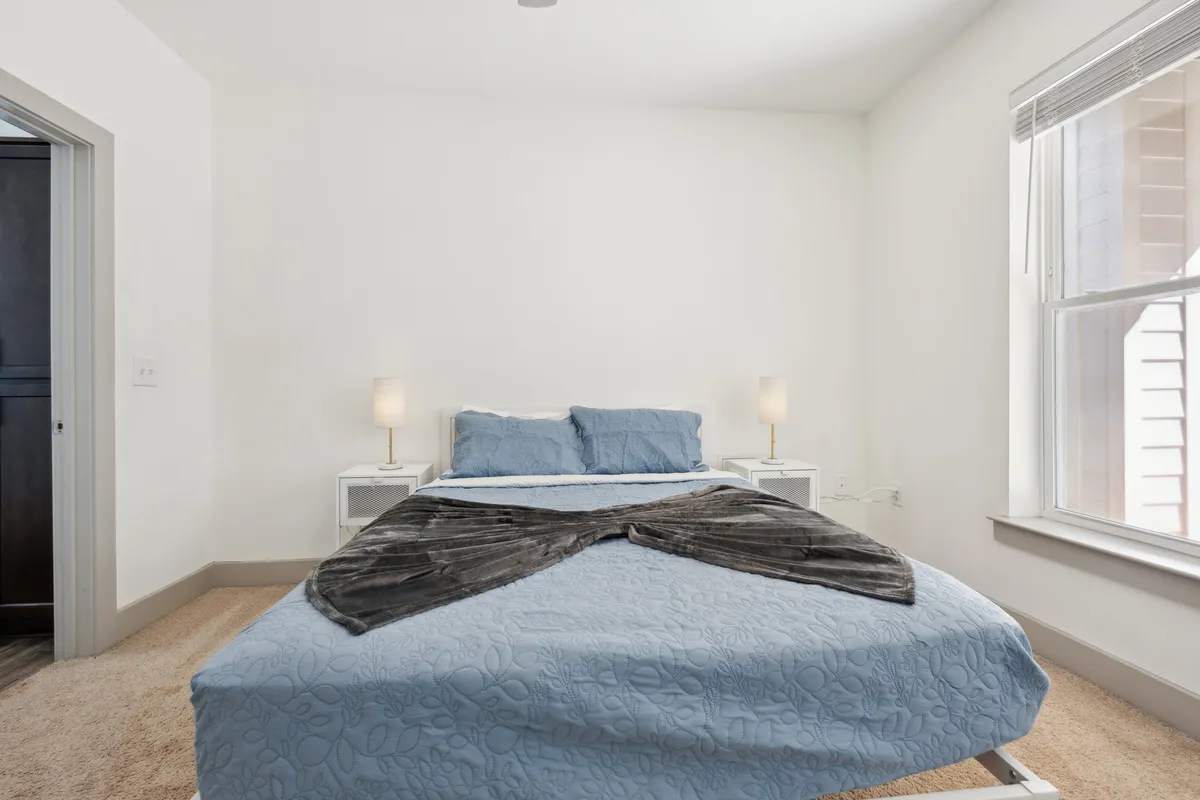A serene bedroom with a bed dressed in blue bedding, nightstands topped with lamps on either side, and a window letting in natural light. The space feels calm, inviting, and perfectly suited for restful solitude or a cozy moment with a loved one.