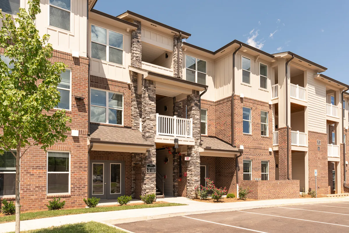 This exterior image showcases the building’s striking brickwork, stone accents, and balconies framed by lush green landscaping. The scene suggests a vibrant residential setting with active social spaces, such as balconies and communal walkways.