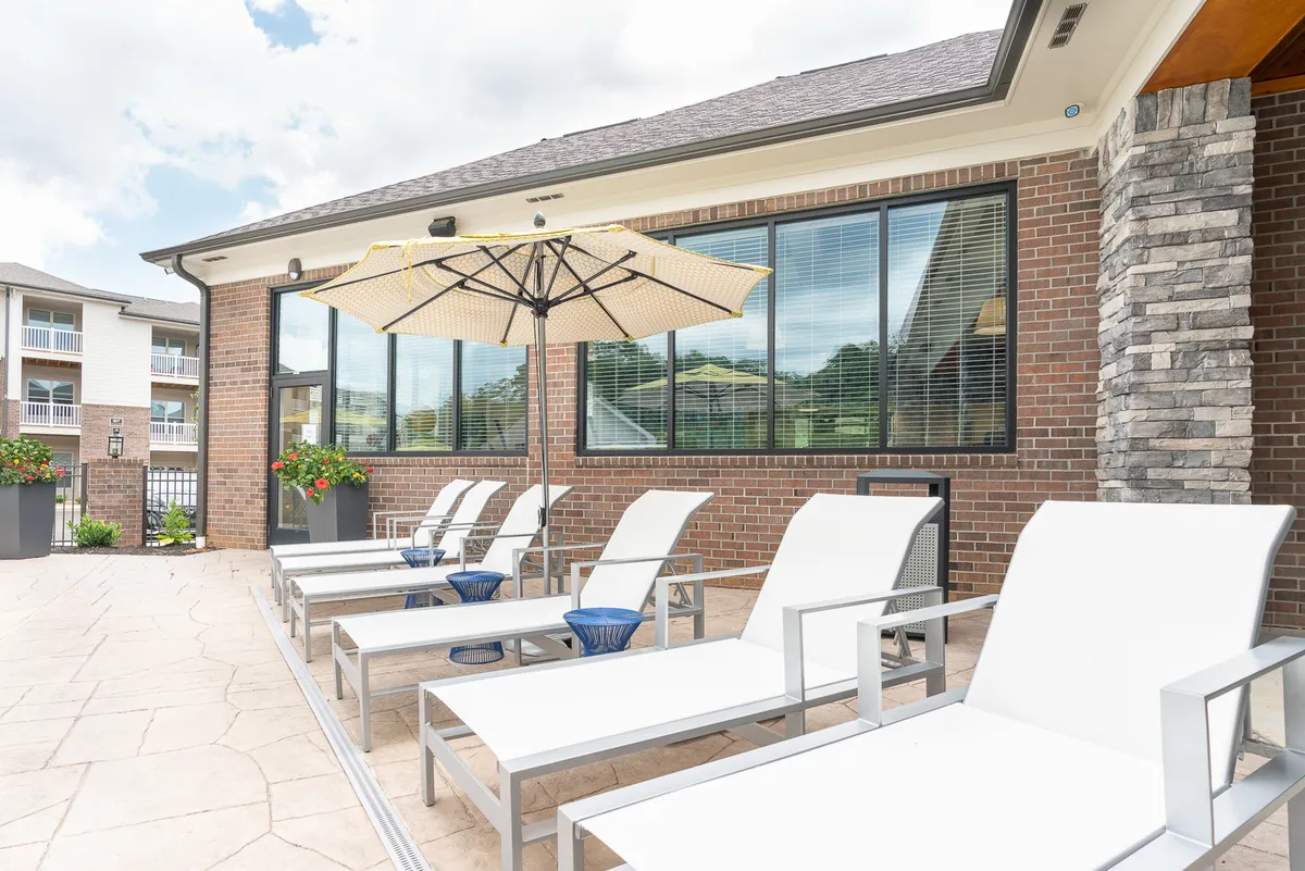 An inviting outdoor lounge area featuring comfortable lounge chairs beneath a large umbrella, set against a backdrop of a brick wall and expansive windows. The area is bathed in natural light, suggesting a perfect spot to unwind after a day of city exploration.