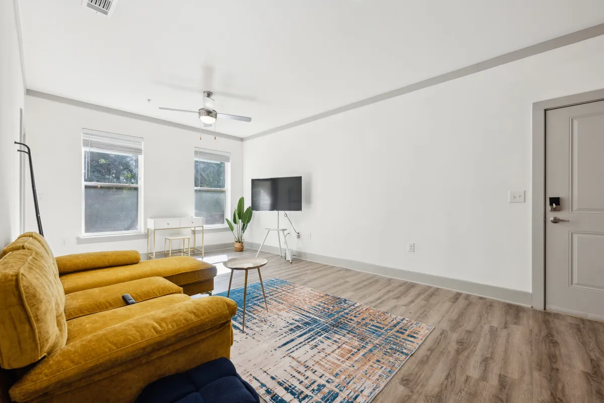 A modern living space featuring a bright yellow chair, a comfortable desk setup with a chair for work, a TV, a plant, and an inviting area rug. The space is well-lit and designed for both productivity (with the desk) and relaxation (with seating and entertainment options).