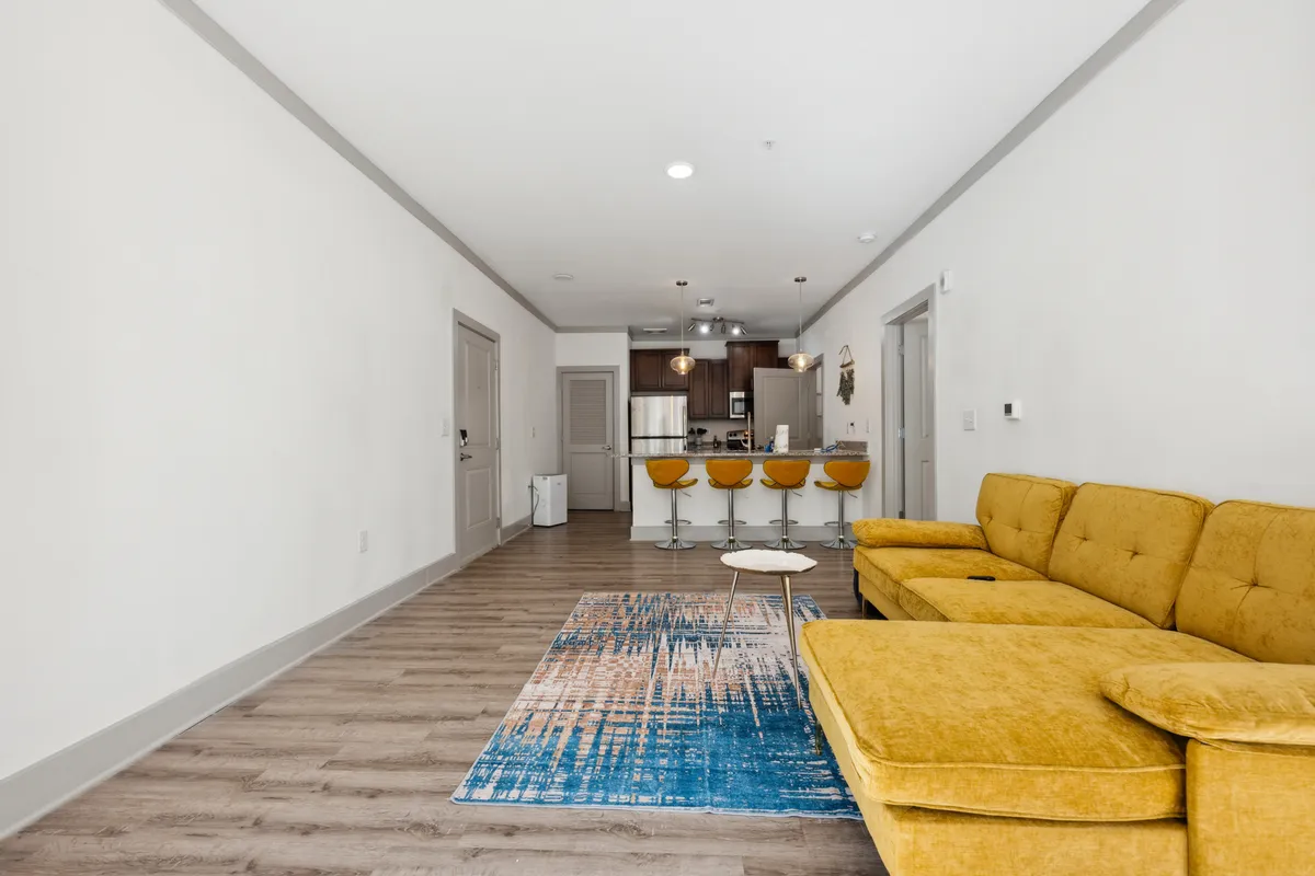 A colorful and lively living area featuring a bright yellow sectional sofa, a blue and orange area rug, and bar stools at the kitchen counter. The furniture arrangement facilitates both casual lounging and entertaining, and the room radiates a welcoming, vibrant mood.