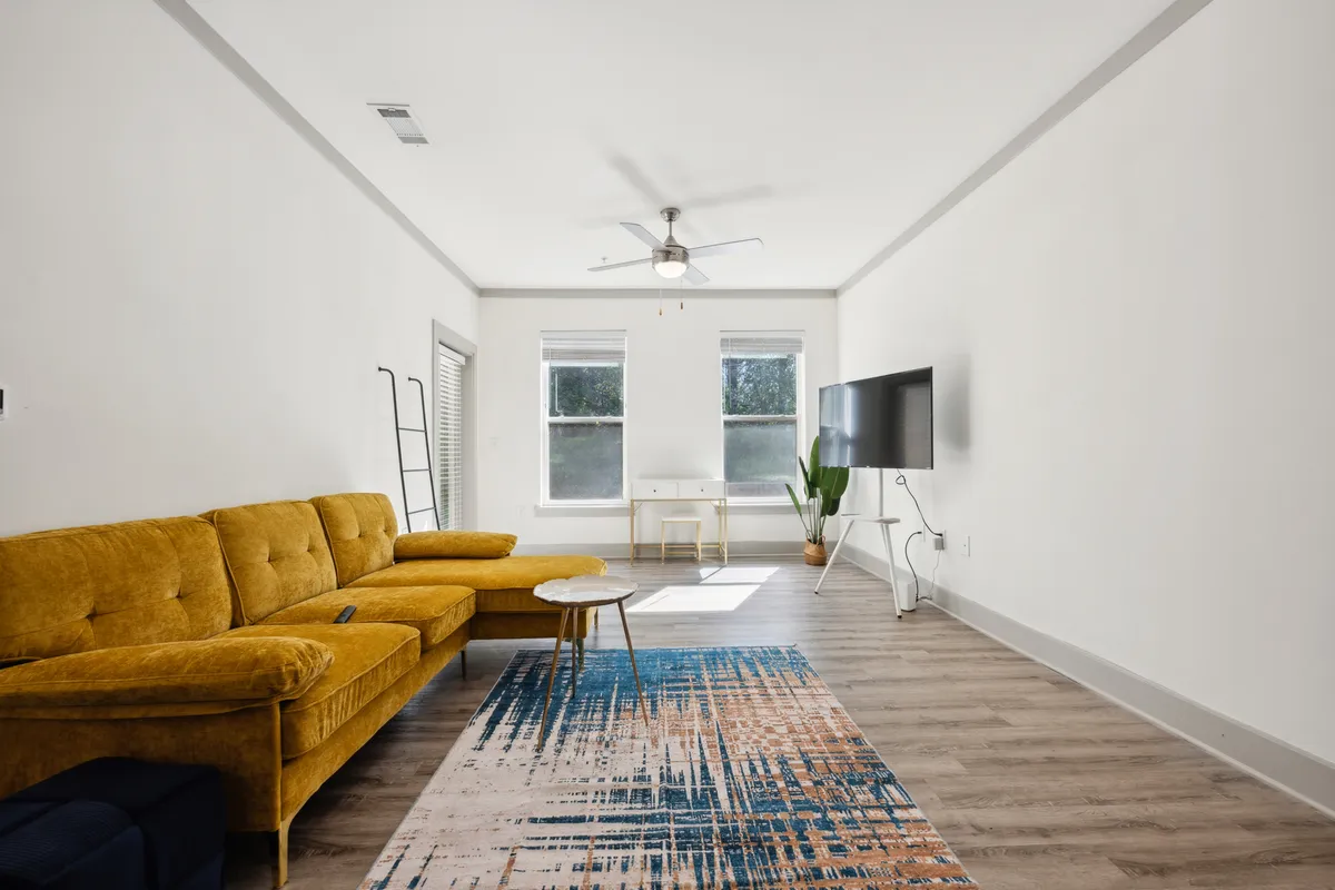 A modern living room with a yellow couch, coffee table, soft rug, television, and ample windows. The layout emphasizes comfort and relaxation, with natural light streaming in, creating a cozy nook for streaming, reading, or quiet afternoons.