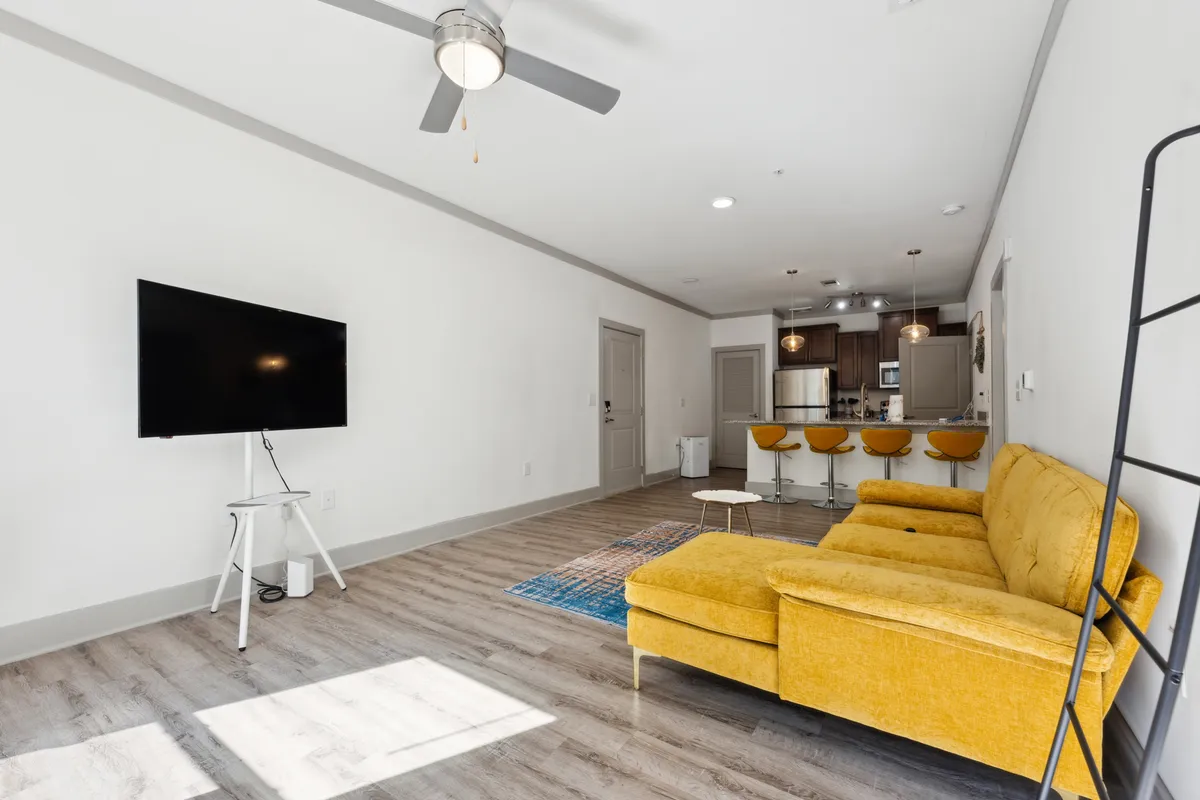 A vibrant, open living area centered around a stylish yellow couch facing a large television. Bar stools delineate the nearby kitchen space, and the room is filled with natural light from generous windows. The space is arranged for both relaxation and entertainment, encouraging socializing and movie nights.
